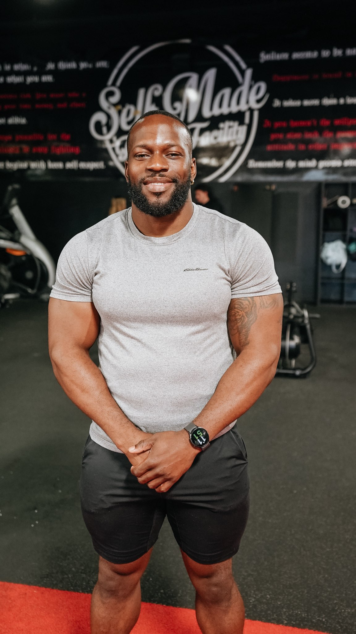 A fit man in his early 30s, wearing workout clothes, smiles confidently in front of a weightlifting platform.